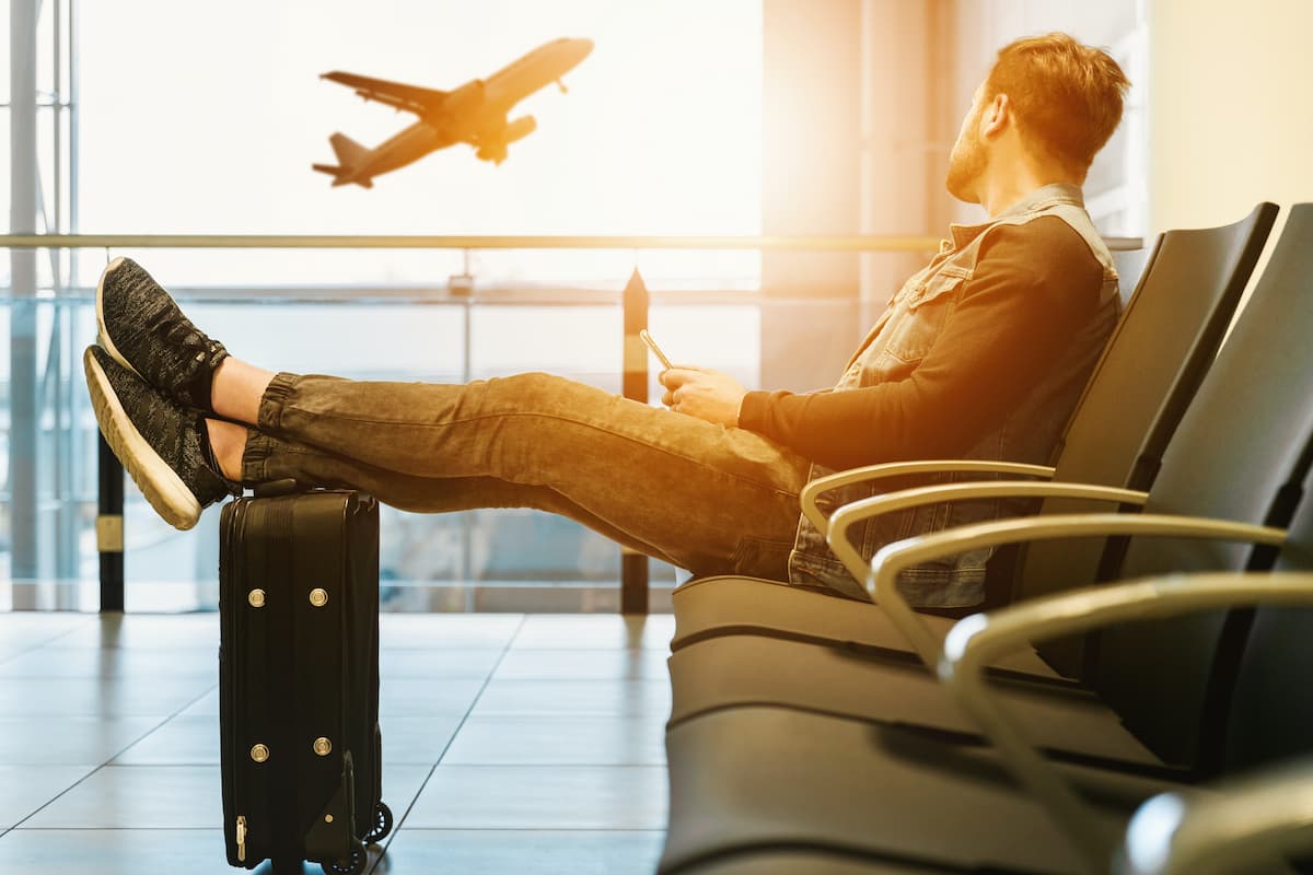 A man is sitting on a gang chair inside the airport, looking at the airplane taking off. 