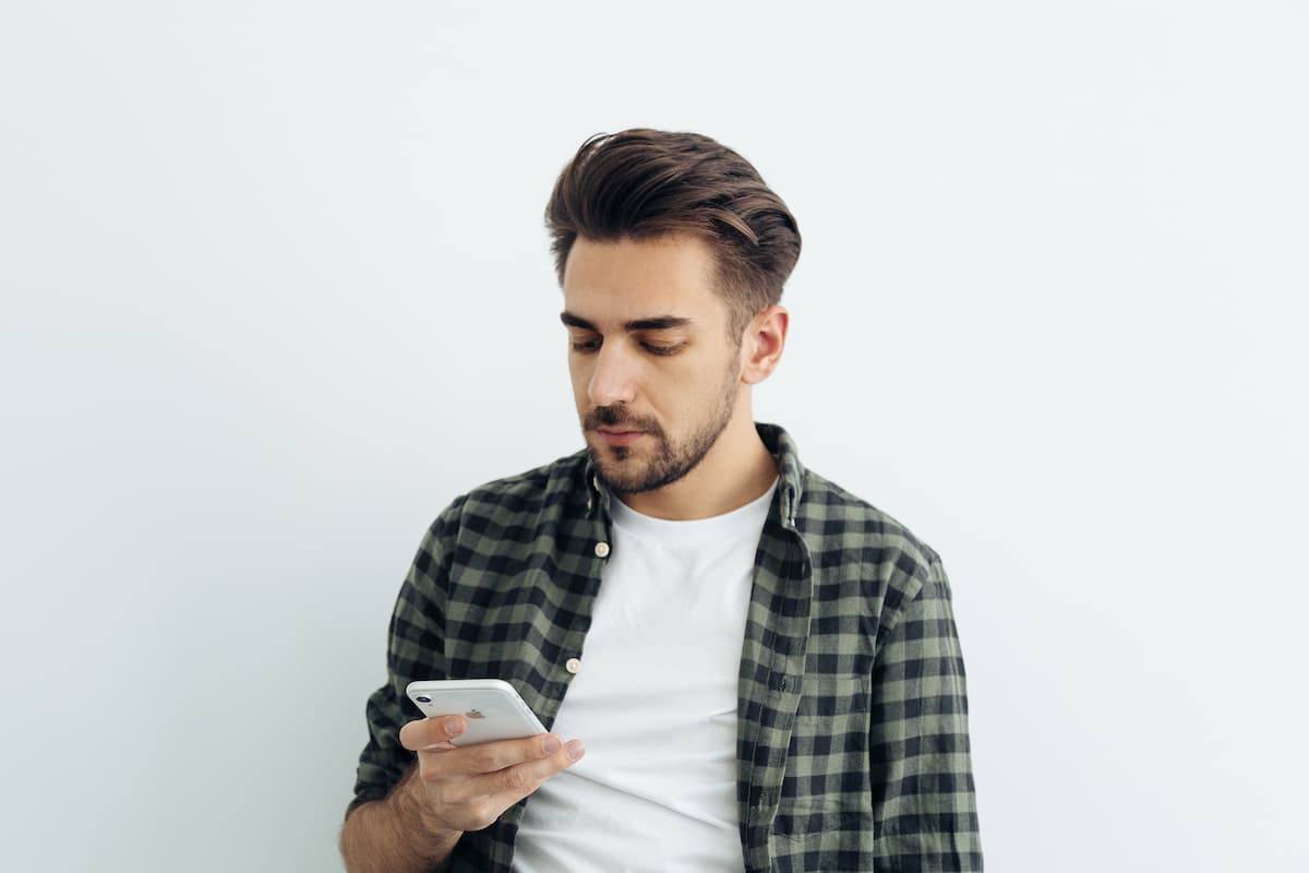 A man in a white t-shirt over a checkered shirt using his iPhone with a white background.