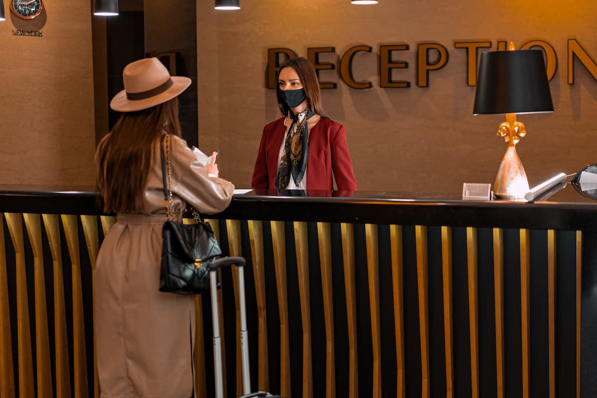 A woman in a brown coat and cowboy hat is talking to the receptionist in the hotel lobby. 