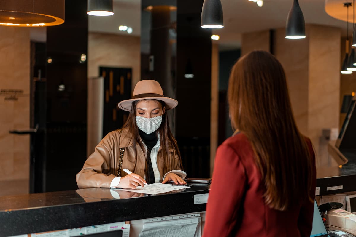 A woman wearing a cowboy hat and a mask is signing a paper at the front desk. 