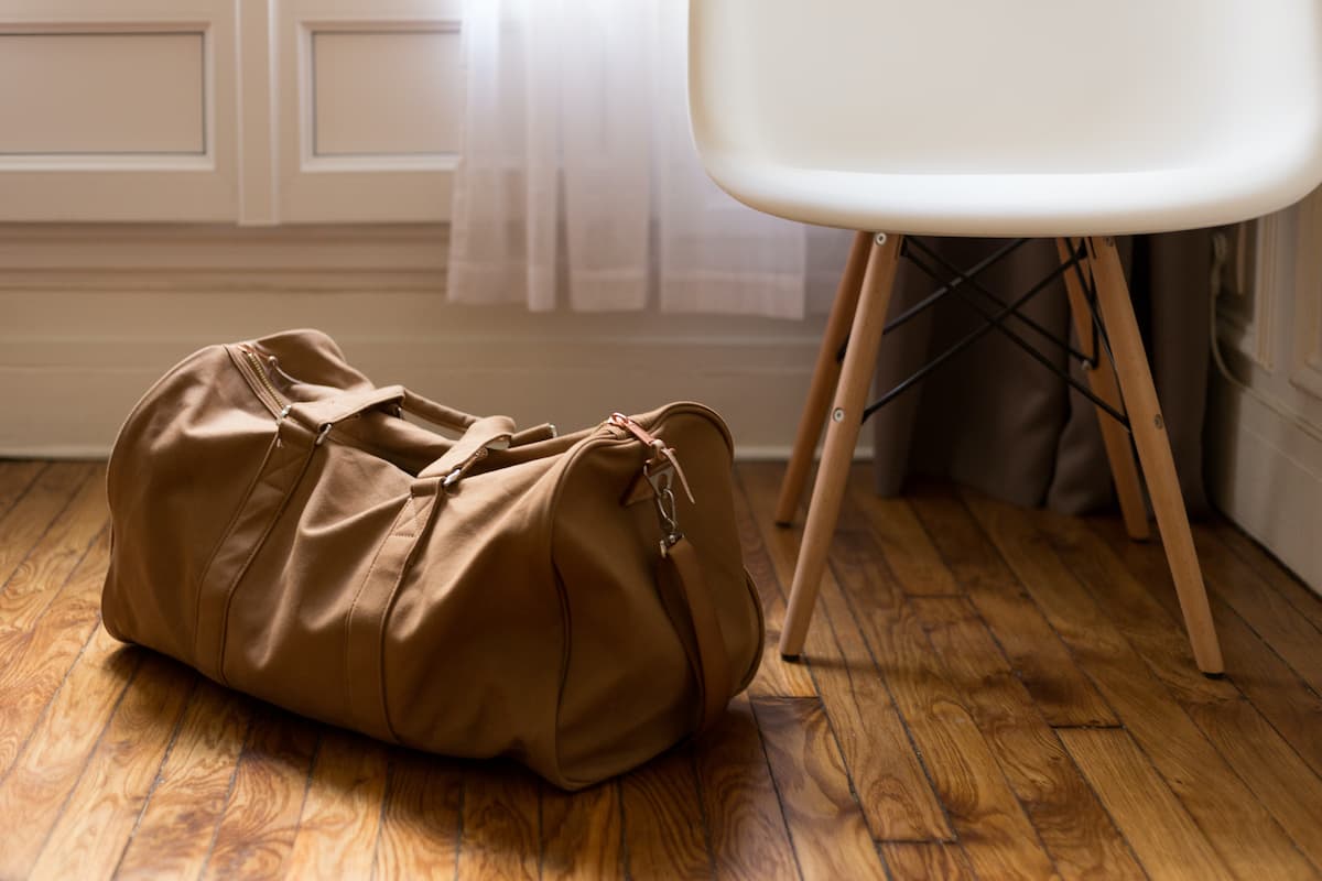 A brown duffel bag is on a wooden floor beside a chair. 