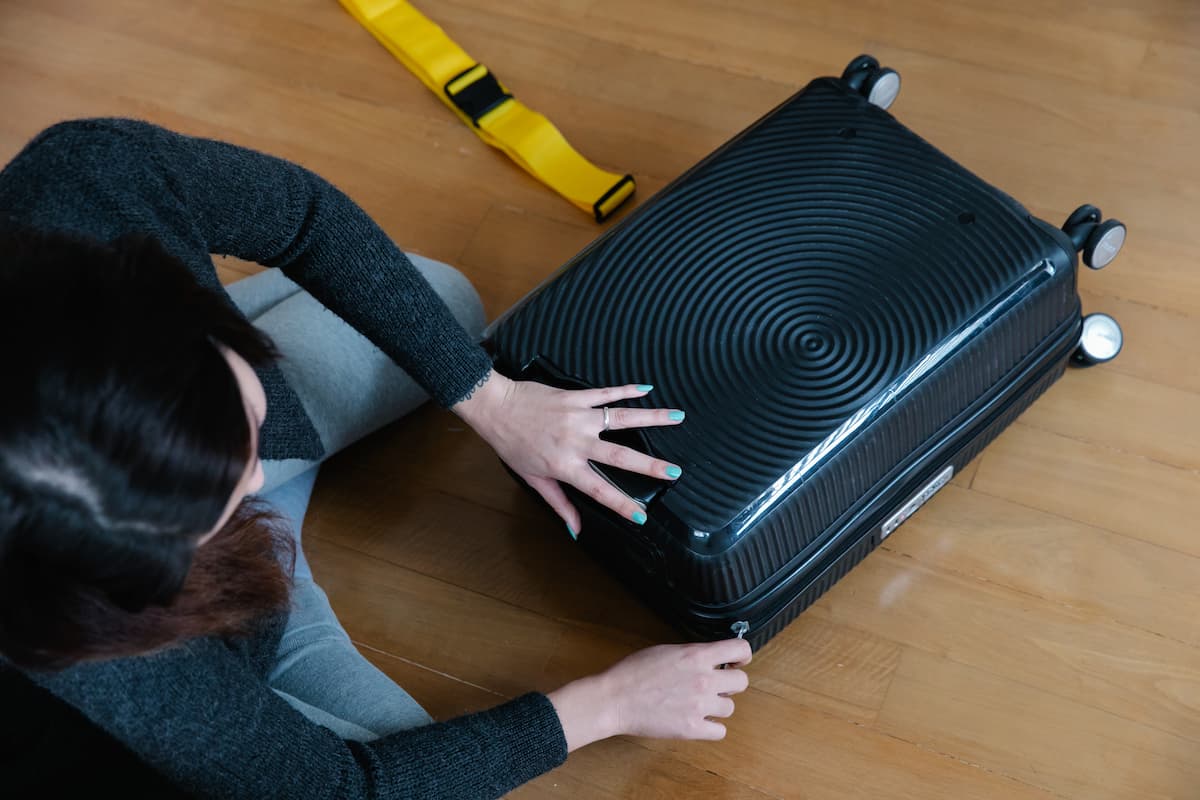 A woman sitting on a wooden floor zipping her black luggage. 