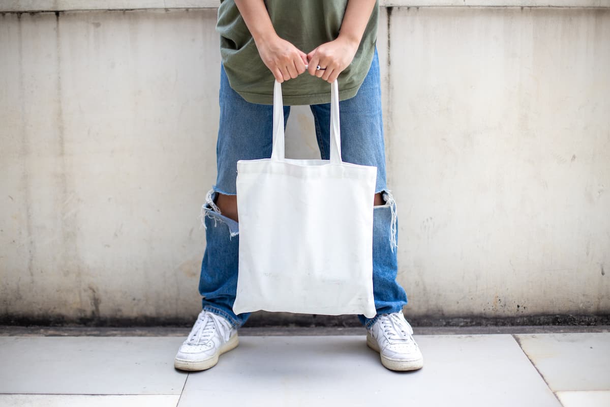 Cropped photo of a man in ripped jeans and white shoes holding a white tote bag. 